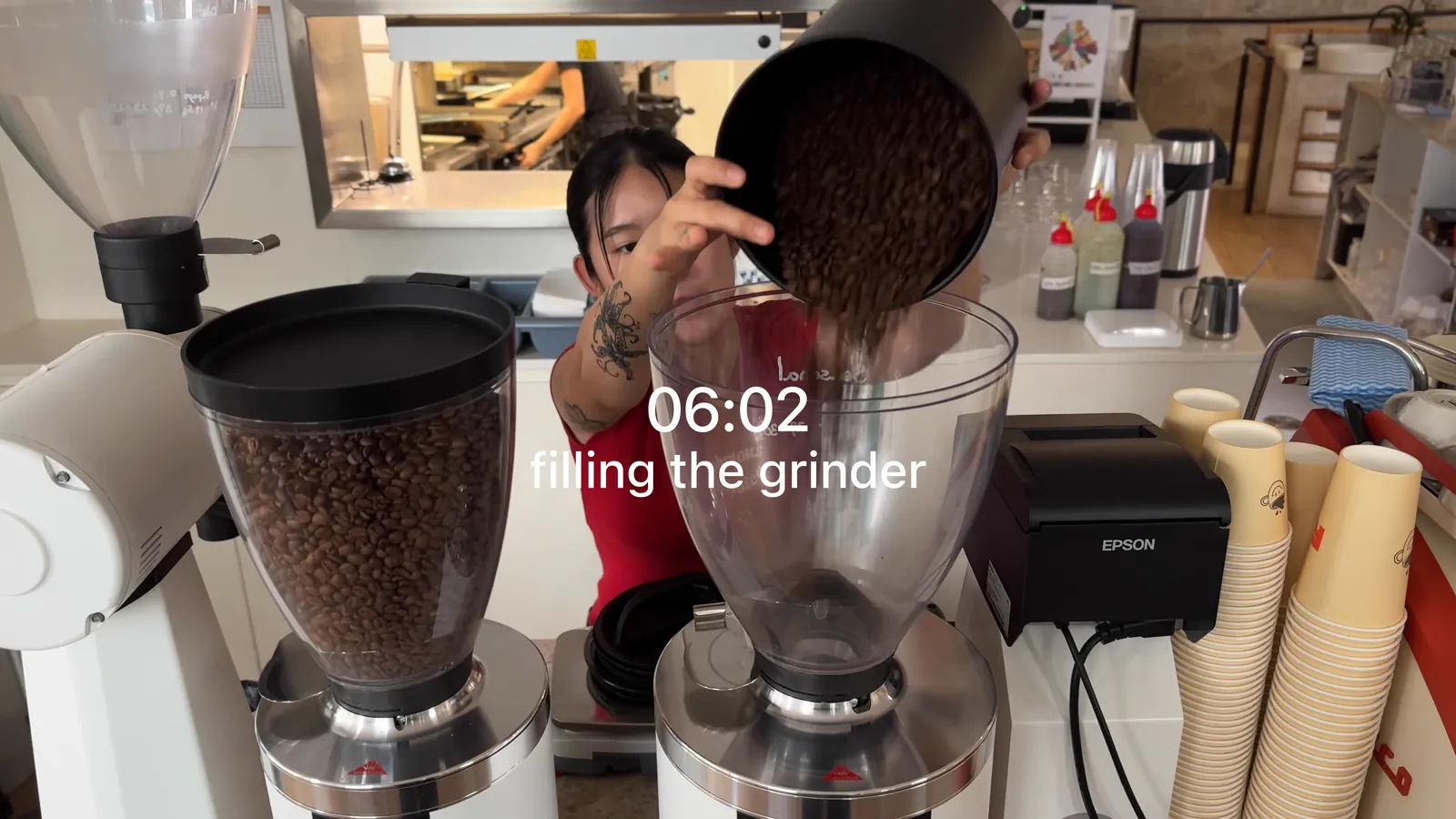 A barista tips coffee beans into a grinder hopper at FAVE Specialty Coffee, with 06:02 and filling the grinder written across the frame
