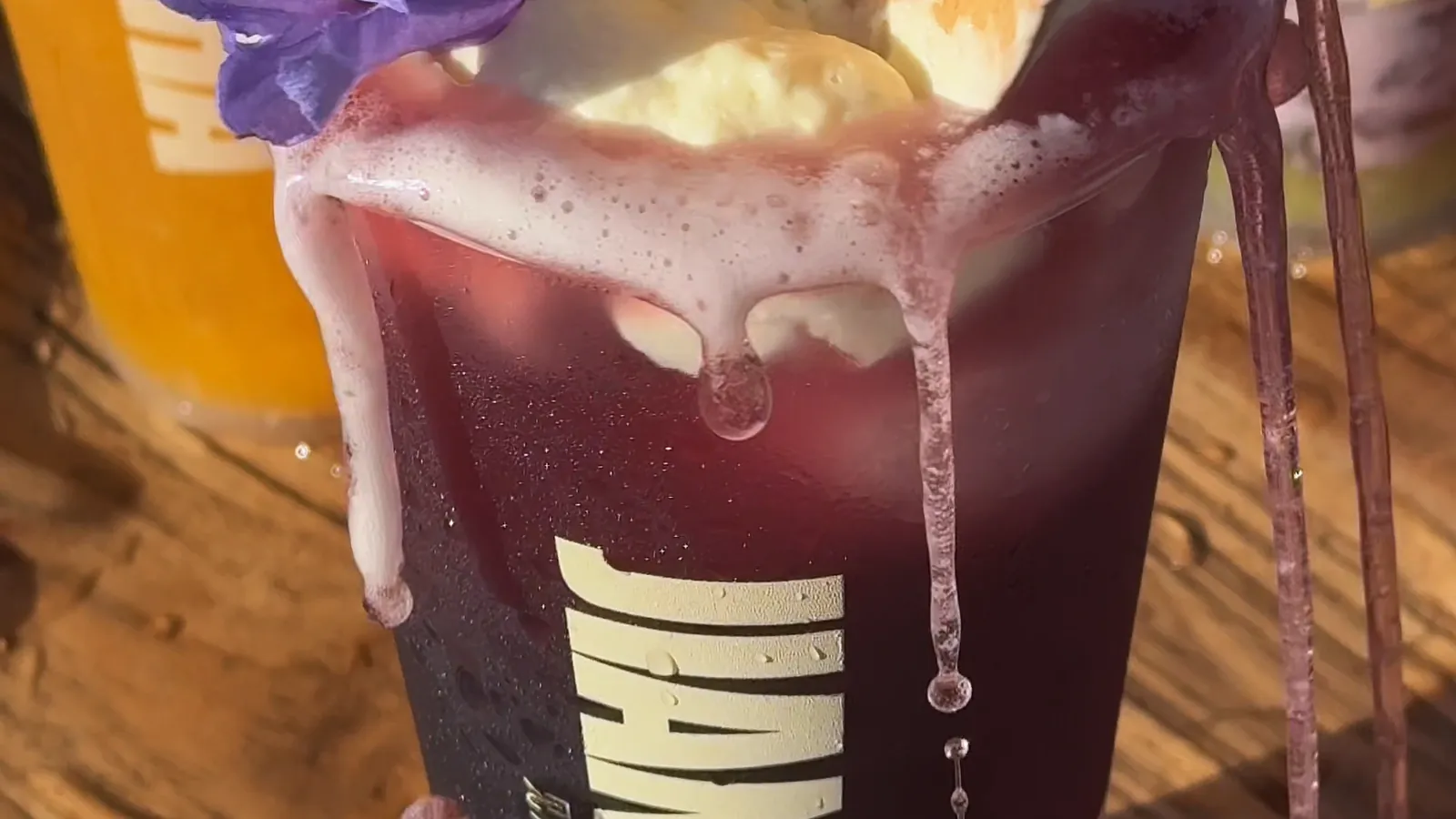 A purple Jian Cha slushie topped with cream cheese and a jacaranda flower, spilling over the rim and down the side of the cup