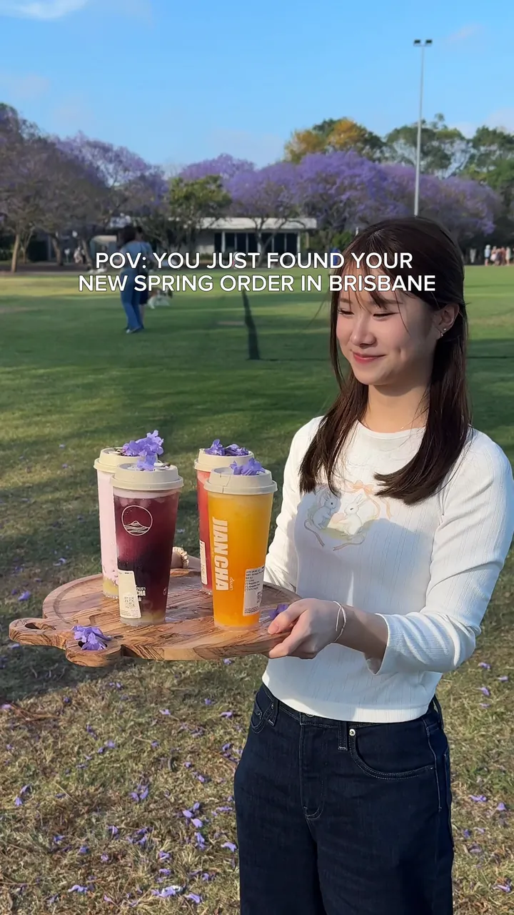 A woman in New Farm Park holding a wooden tray of four Jian Cha drinks, each topped with jacaranda petals