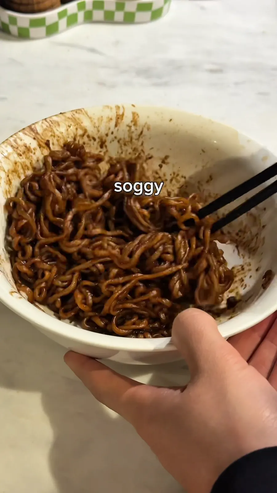 A hand holds a bowl of glossy black bean noodles with chopsticks. A single white word in the middle of the frame reads soggy.