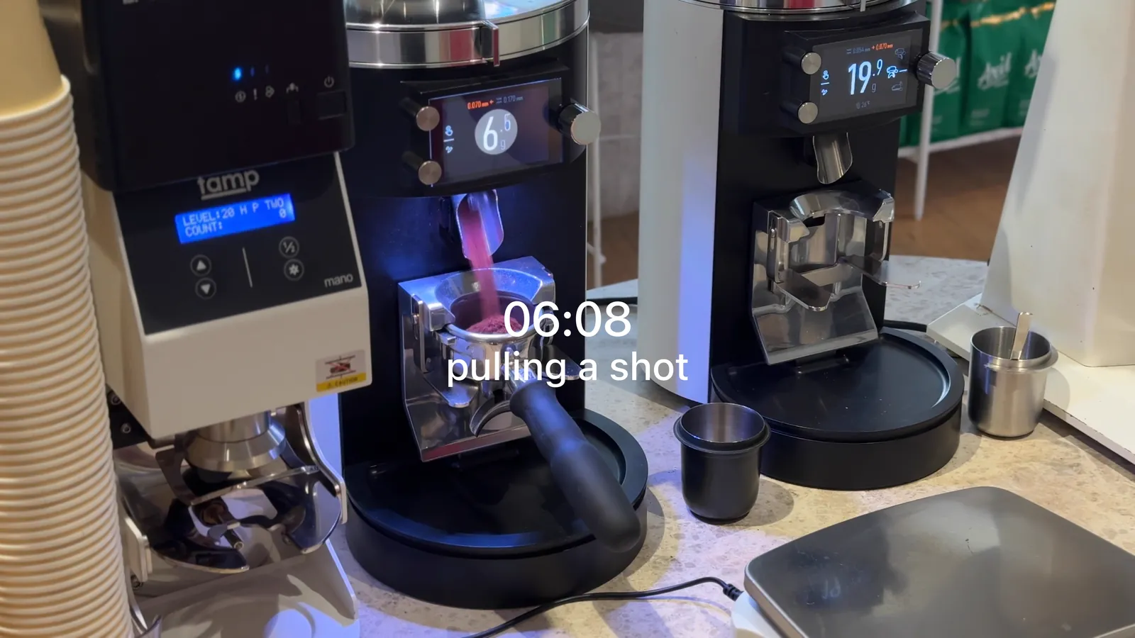 Coffee grinders on the bench at FAVE, one dosing into a portafilter under a pink light. Plain white text in the middle of the frame reads 06:08, pulling a shot.