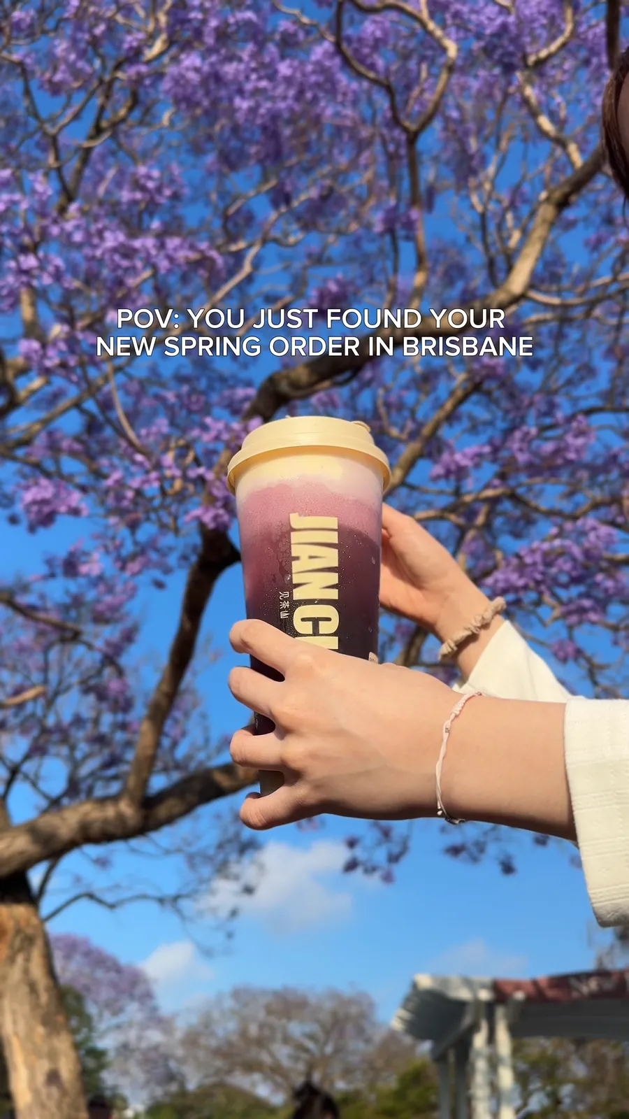 A hand holds a purple Jian Cha slushie up against jacaranda flowers and blue sky. Small white capitals in the top third read POV: you just found your new spring order in Brisbane.