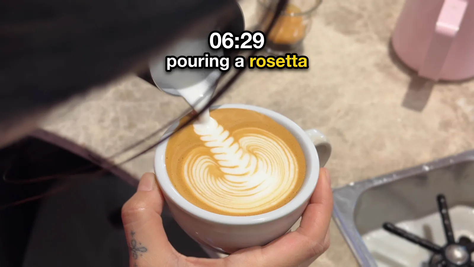 Mock-up: a rosetta being poured into a white cup, with bold white text reading 06:29, pouring a rosetta. The text has a thick black outline and a drop shadow, and the word rosetta is yellow.