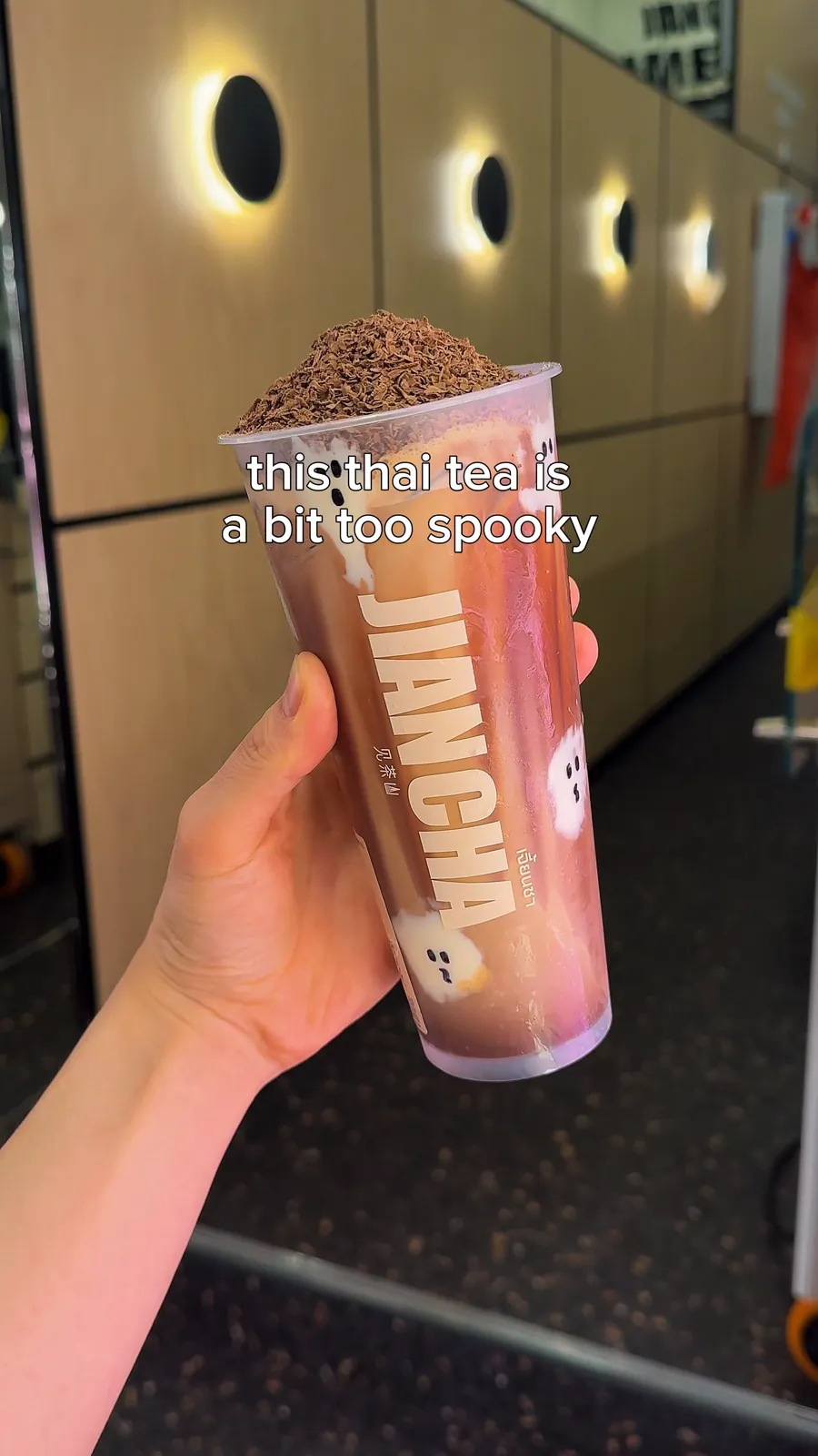 A hand holds up a Jian Cha Thai Milk Tea Cocoa topped with grated chocolate, with mochi ghosts inside the cup. White lowercase text with a thin dark outline reads this thai tea is a bit too spooky.