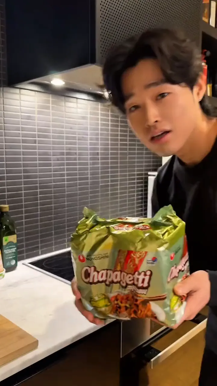 MJ holds up a pack of Chapagetti in a home kitchen, then lifts the noodles and takes a bite.
