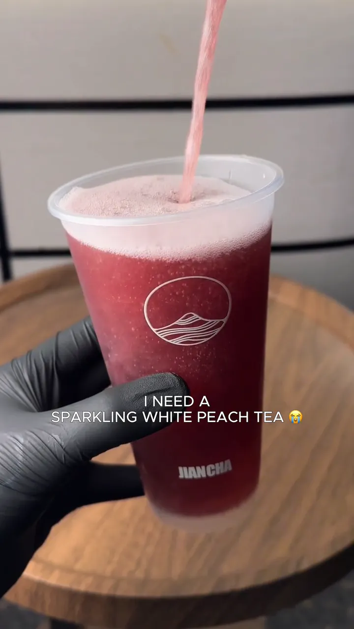 Pink sparkling tea poured into a deep purple drink in a Jian Cha cup, held in a black glove. White text on screen reads “I need a sparkling white peach tea”.